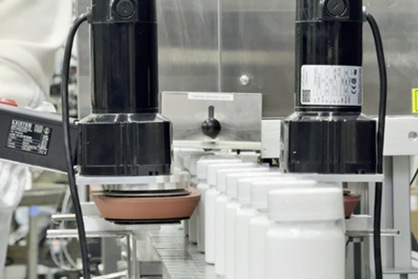 White plastic bottles are conveyed from a Pharmafill tamper-evident neck bander to a Pharmafill heat tunnel